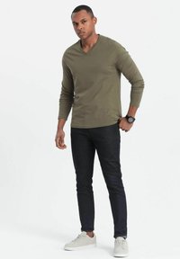 Camisa de manga larga verde oliva con escote en V, combinada con jeans oscuros y zapatillas grises claro. Cuenta con un diseño ajustado y un estilo minimalista.