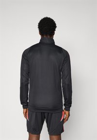 Chaqueta deportiva negra con un cuello alto, mangas largas y textura suave, combinada con pantalones cortos oscuros con acentos rojos.