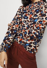 Halenka s dlouhým rukávem s krémovým pozadím, zdobená barevným leopardeím vzorem v hnědé, červené a modré barvě. Volánkový výstřih a manžety.