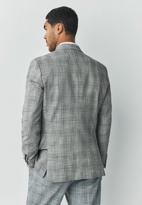 Chaqueta de traje a cuadros grises con cierre de dos botones, aberturas laterales y costuras detalladas. La tela tiene un patrón texturizado sutil.