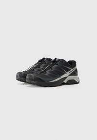 Black trail running shoes with a textured upper, molded toe cap, and elastic quick-lacing system. Features a grey accent along the side.