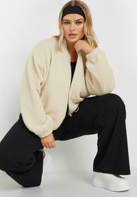 Kremowy, puszysty bomber z polaru z zapięciem na zamek. Opadające ramiona i elastyczne mankiety. Noszony z czarnymi spodniami i białymi trampkami.