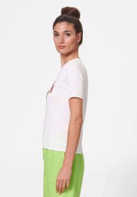 White short-sleeve t-shirt with a round neckline and a colorful graphic design. Model wears lime green pants, showcasing a relaxed fit and cotton material.
