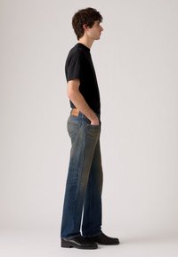 Dark blue denim jeans with a gradual fade, straight cut, front pockets, and a leather patch at the back waistband. Paired with a black t-shirt and shoes.