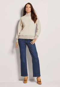 Beige coltrui met ribbelzoom en -manchetten, gecombineerd met donkerblauwe flared jeans en bruine platform schoenen. Glad stoffen textuur.