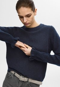 Temno modra pulover iz mehkega pletenega materiala z okroglim izrezom in nabranimi manšetami. Nosim se z svetlo sivim pasom in temnimi kavbojkami.