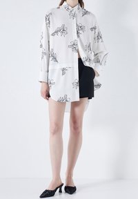 Camicia bianca con bottoni, caratterizzata da motivi floreali neri, maniche lunghe, vestibilità rilassata e orlo curvo. Abbinata a pantaloni corti neri e mules.