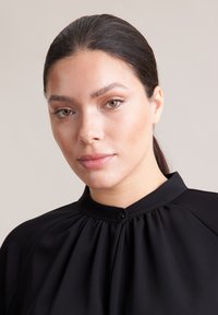Schwarze Bluse mit hohem Kragen, plissiertem Design und einem kleinen Knopfdetail am Hals. Glatte Textur und taillierte Passform.
