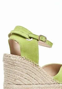 Sandale compensée en suède vert avec une sangle cheville et une boucle métallique, présentant une semelle plateforme en jute texturé.