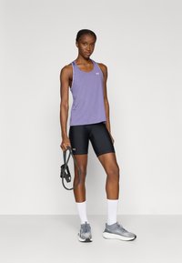 Μωβ αθλητικό τοπ με σχεδιασμό racerback, μαύρο σορτς ψηλόμεσο, γκρι παπούτσια για τρέξιμο και κρατώντας μια λάστιχο αντίστασης.