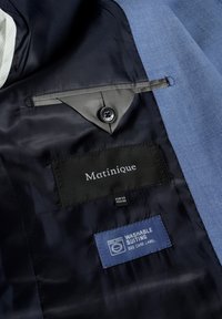 Mėlynos spalvos kostiumo vidus, kuriame yra juodas etiketė su užrašu "Matinique", priežiūros etiketė, nurodanti "PLAUKIAMA KOSTIUMAS" ir mygtuko detalė.