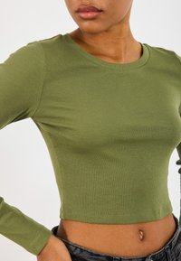 Olíva zöld, bordázott, hosszú ujjú crop top kerek nyakkal és testhezálló fazonnal, sötét farmernadrággal párosítva.