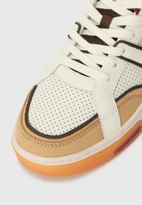 Witte geperforeerde leren sneaker en bruine suède met crèmekleurige veters, met een oranje rubberen zool en bruine accenten op de bovenkant.