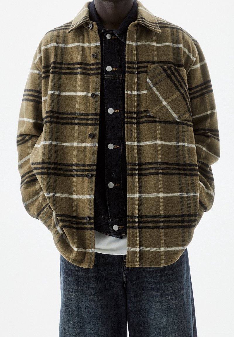 Camisa chaqueta de cuadros marrones y negros en material suave, con un bolsillo en el pecho y cierre de botones. Lleva puesta sobre una capa de denim oscuro.