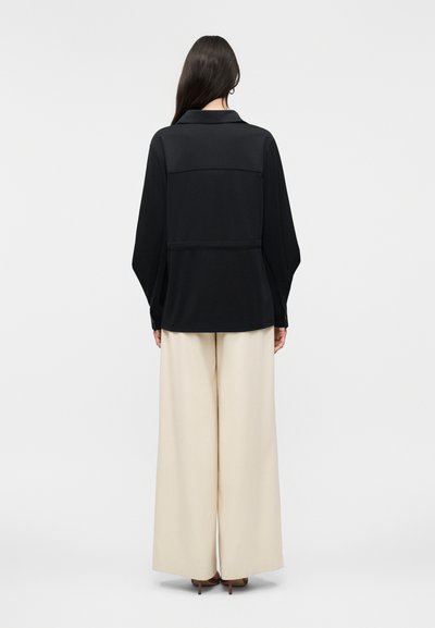 Γυναίκα με μακριά σκούρα μαλλιά, φοράει μαύρο πουκάμισο με μακριά μανίκια και μπεζ παντελόνι φαρδιάς γραμμής, στέκεται με την πλάτη γυρισμένη σε απλό φόντο.