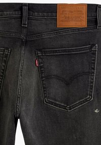 Los vaqueros de mezclilla negros presentan un acabado texturizado, un parche de cuero marrón en la cintura y una distintiva etiqueta roja en el bolsillo trasero.