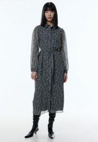 Schwarzes Blumenkleid mit einem knöpfbaren Kragen, langen durchsichtigen Ärmeln und einem Gürtel in der Taille. Kombiniert mit knielangen, glänzend schwarzen Stiefeln.