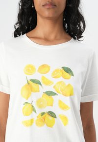Weißes T-Shirt mit einem bunten Druck von ganzen und geschnittenen Zitronen sowie grünen Blättern, das ein detailliertes Design in lebendigen Gelbtönen zeigt.