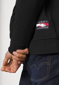 Czarna bluza z prążkowanym dołem, z kolorową naszywką z logo przedstawiającą dłonie. W zestawie z ciemnymi jeansami z wyraźnym przeszyciem.