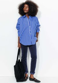 Chemise rayée bleue et blanche avec une poche poitrine, accompagnée d'un pantalon marine. Sac fourre-tout noir et chaussures à enfiler avec des accents cloutés.