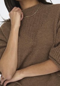 Hnědý pletený svetr s vysokým límcem, který má texturovaný vzor a volný střih. Zlatý řetízek zvýrazňuje výstřih.