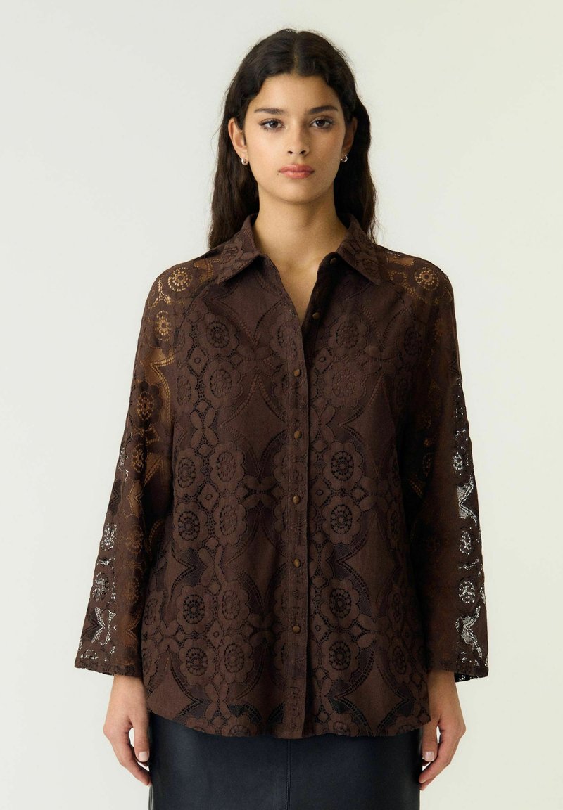 Jeune femme portant un chemisier en dentelle marron foncé avec boutons, motifs floraux et manches longues, debout devant un fond clair uni.