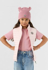 Roze gewatteerd vest met een hoge kraag over een roze T-shirt, gecombineerd met een lichtroze gebreide muts met ronde oren. Blauwe denim shorts.