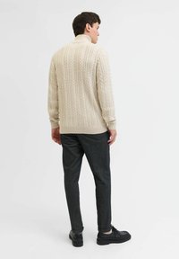 Beżowy sweter z dzianiny z wysokim kołnierzem, teksturowanym wzorem i ściągaczami przy mankietach, w zestawie z ciemnoszarymi spodniami o dopasowanym kroju i czarnymi butami.