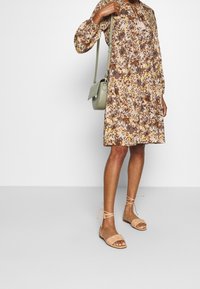 Kukallinen mekko vaaleassa beigeessä, ruskeassa ja keltaisessa, pitkillä hihoilla, yhdistettynä beigeihin nauhatai sandaaleihin ja vaaleanvihreään olkalaukkuun.