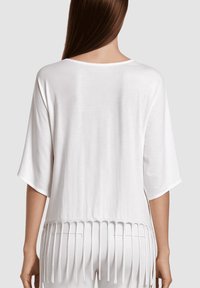 Mujer con una blusa blanca suelta de mangas hasta el codo y detalles de flecos verticales en el dobladillo, vista desde la espalda.