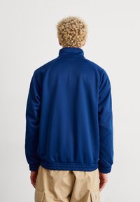 Mies, jolla on kiharat vaaleat hiukset, pukeutuneena siniseen pitkähihaiseen takkiin ja vaaleisiin cargo-shorteihin, nähtynä takaa.