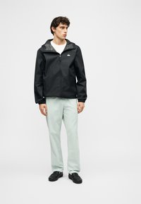 Νεαρός άνδρας φοράει μαύρο φούτερ με φερμουάρ και κουκούλα, λευκό πουκάμισο, ανοιχτόχρωμο παντελόνι και μαύρα παπούτσια, στέκεται μπροστά σε απλό φόντο.