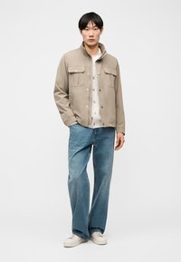 Homme debout portant une veste beige, une chemise blanche boutonnée, un jean bleu et des baskets blanches sur fond blanc uni.