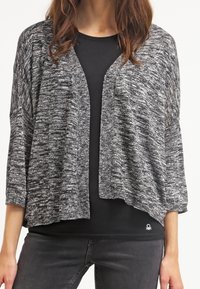 Mujer vestida con un cárdigan de punto negro y gris, abierto por delante, sobre una camiseta negra y pantalones gris oscuro, de pie con las manos relajadas a los lados.