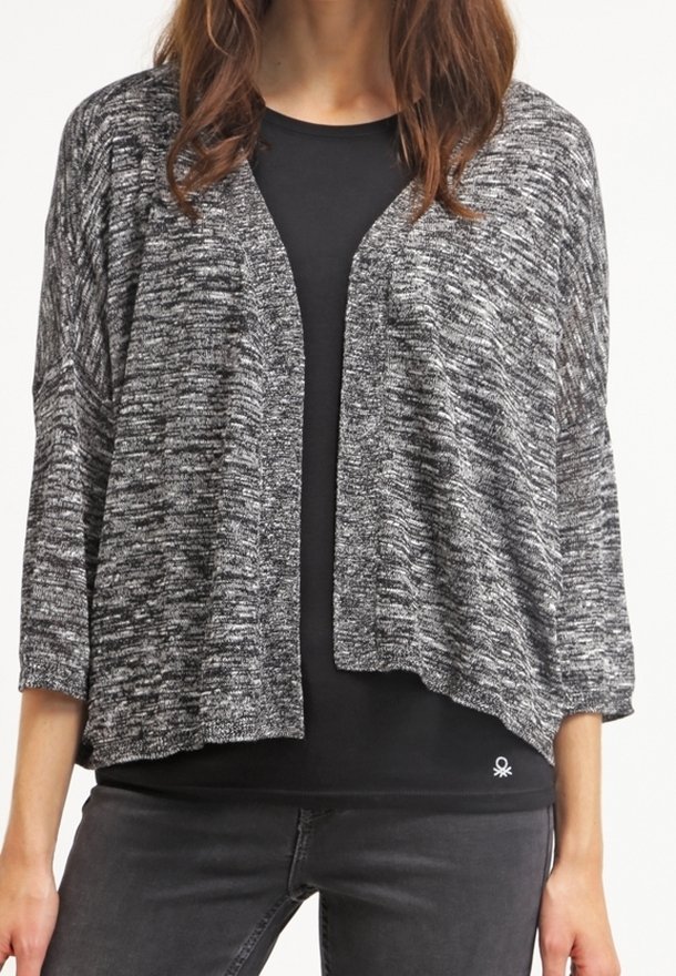 Mujer vestida con un cárdigan de punto negro y gris, abierto por delante, sobre una camiseta negra y pantalones gris oscuro, de pie con las manos relajadas a los lados.