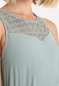 Ljusgrön ärmlös topp med en dekorativ spetspanel vid halsringningen och ett mjukt, flödande tyg med en avkopplad passform.