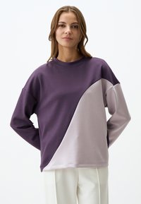 Felpa viola e grigia chiara con un design a spacco diagonale, texture morbida, colletto a girocollo e vestibilità rilassata. Impostata su uno sfondo semplice.