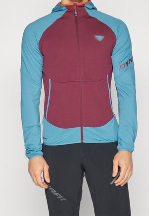 Mann trägt eine enganliegende Kapuzenjacke mit bordeauxroter Front und blauen Ärmeln und Kapuze, kombiniert mit schwarzen Hosen mit grauem Schriftzug.