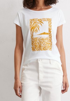 Woman wearing white T-shirt with yellow graphic of palm leaves, waves, and a distant island, paired with high-waisted white pants.