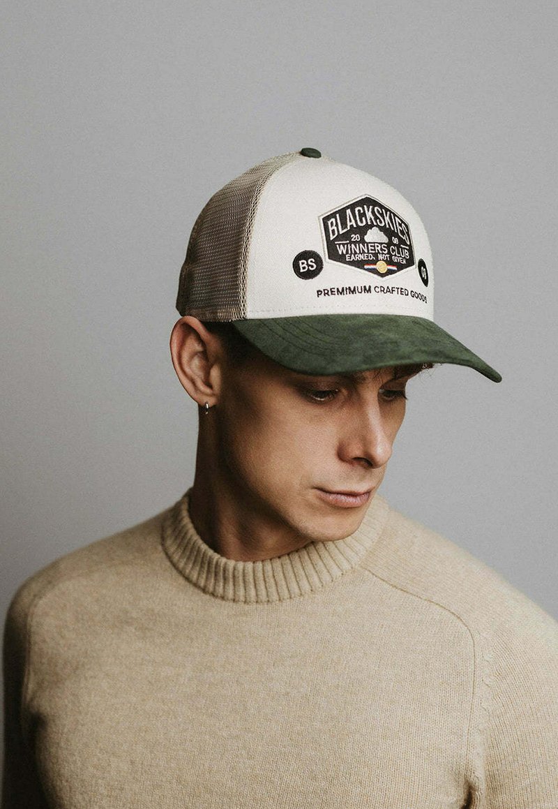 Joven con suéter beige y gorra Blackskies beige y verde, mirando hacia abajo y hacia un lado sobre un fondo gris liso.