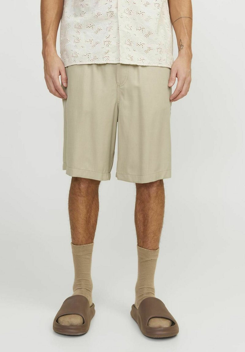 Shorts beiges avec une coupe droite et des poches latérales, associés à des chaussettes beiges et des sandales à glisser marron. Porté avec une chemise légère à motifs.