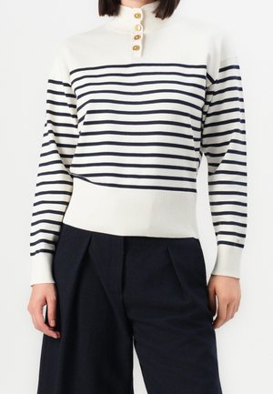 Femme portant un pull blanc à rayures horizontales noires, un col montant boutonné, et un pantalon large noir taille haute.