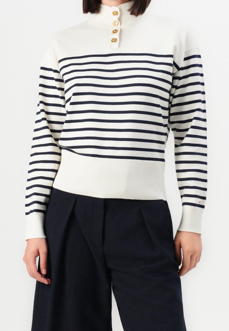 Femme portant un pull blanc à rayures horizontales noires, un col montant boutonné, et un pantalon large noir taille haute.