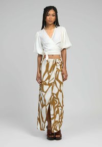 Haut enroulé blanc à manches bouffantes associé à une longue jupe à motif abstrait brun et blanc avec une fente sur le côté. Le mannequin porte des sandales compensées brunes.