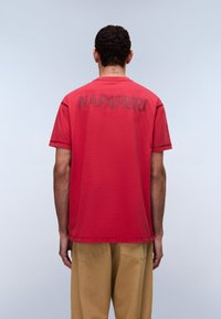 Κόκκινο βαμβακερό t-shirt με ανάγλυφο κείμενο "NAPAPIRRI" στην πλάτη, στρογγυλή λαιμόκοψη και κοντά μανίκια. Φορεμένο με μπεζ παντελόνια.