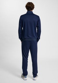 Tuta di colore blu navy con colletto alto e polsini elasticizzati, caratterizzata da una texture liscia e gambe dei pantaloni affusolate, abbinata a scarpe grigie.