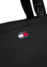 Czarna torba tote z nylonu o prostokątnym kształcie, z naszywką z logo TOMMY JEANS w niebieskim, czerwonym i białym kolorze. Grube czarne paski.