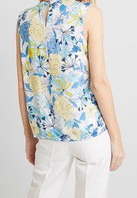 Top sans manches floral avec un motif bleu, jaune et blanc, doté d'un détail au dos en forme de trou de serrure, fabriqué dans un matériau léger et lisse.