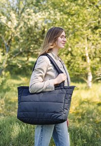 Svart puffig handväska med två stadiga axelremmar, rektangulär form och sydd textur, buren av en person i en naturlig miljö.