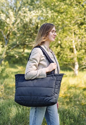 Svart puffig handväska med två stadiga axelremmar, rektangulär form och sydd textur, buren av en person i en naturlig miljö.